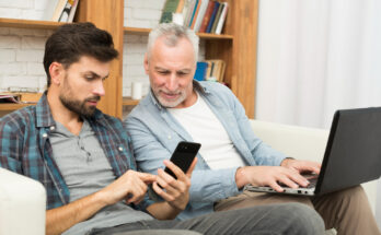 Un jeune homme et un homme senior sont assis côte à côte sur un canapé, échangeant avec complicité autour d'un smartphone et d'un ordinateur portable
