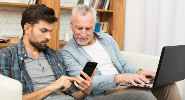 Un jeune homme et un homme senior sont assis côte à côte sur un canapé, échangeant avec complicité autour d'un smartphone et d'un ordinateur portable
