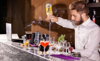 Un barman barbu en chemise blanche verse un ingrédient à l'envers depuis une bouteille pour préparer un cocktail sur un comptoir en marbre où s'élève de la fumée.
