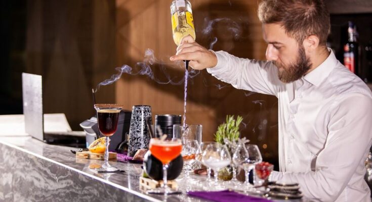 Un barman barbu en chemise blanche verse un ingrédient à l'envers depuis une bouteille pour préparer un cocktail sur un comptoir en marbre où s'élève de la fumée.