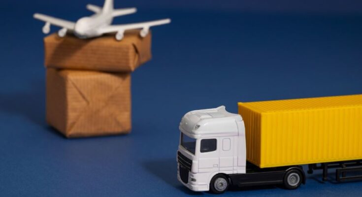 Une miniature de camion blanc à remorque jaune et un petit avion blanc posé sur des paquets en carton, symbolisant la logistique et le transport international sur fond bleu.
