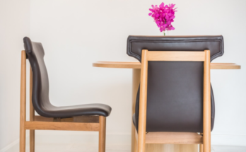 Deux chaises en bois clair avec un revêtement en cuir sombre entourent une table ronde sur laquelle est posé un petit vase de fleurs fuchsia