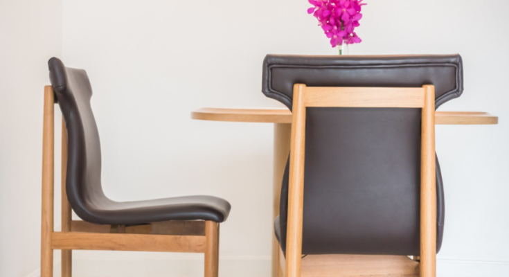 Deux chaises en bois clair avec un revêtement en cuir sombre entourent une table ronde sur laquelle est posé un petit vase de fleurs fuchsia