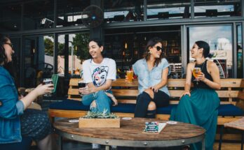 un groupe de quatre amies souriantes partageant des verres et discutant de manière conviviale sur la terrasse d'un bar moderne.