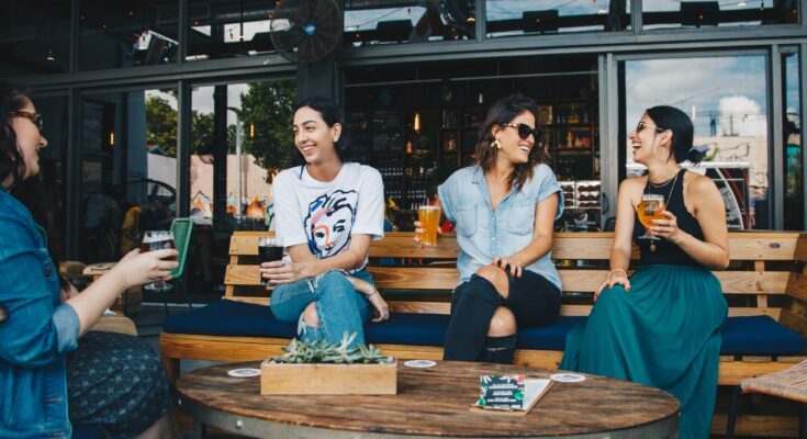 un groupe de quatre amies souriantes partageant des verres et discutant de manière conviviale sur la terrasse d'un bar moderne.