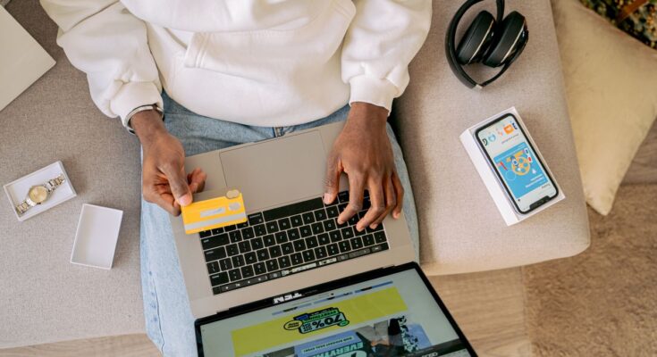 une personne vue du dessus, installée sur un canapé, utilisant sa carte bancaire et son ordinateur portable pour effectuer des achats en ligne