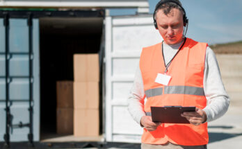 un agent logistique équipé d'un casque micro et d'un gilet haute visibilité consultant un porte-bloc devant un conteneur de fret ouvert.