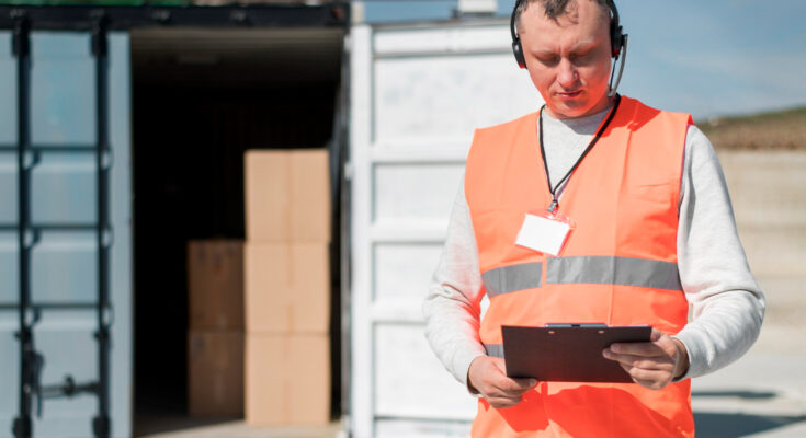 un agent logistique équipé d'un casque micro et d'un gilet haute visibilité consultant un porte-bloc devant un conteneur de fret ouvert.