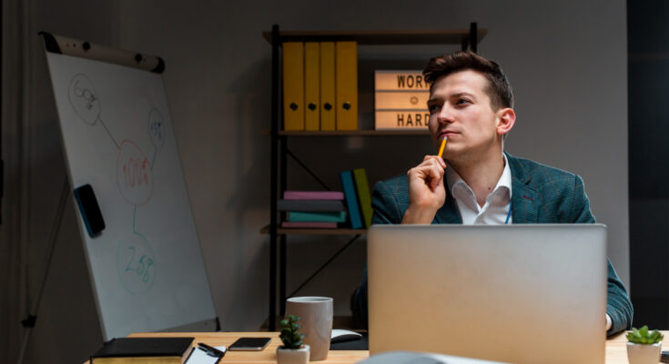 un jeune entrepreneur ou professionnel pensif à son bureau, le crayon posé sur le menton en pleine réflexion
