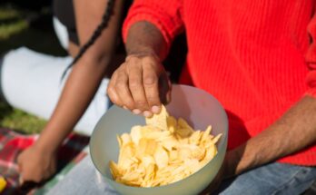une personne portant un pull rouge qui pioche une chip ondulée dans un grand bol en plastique rempli de chips