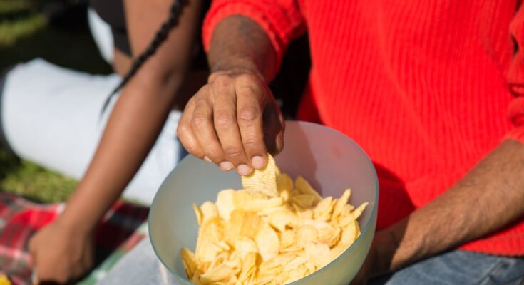 une personne portant un pull rouge qui pioche une chip ondulée dans un grand bol en plastique rempli de chips