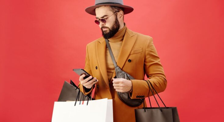 une veste marron et d'un chapeau gris, consultant son smartphone tout en portant plusieurs sacs de shopping