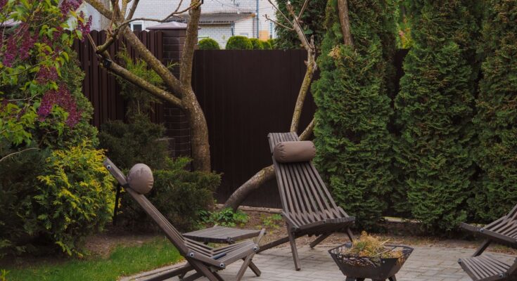 un jardin bordé de verdure, composé de chaises longues en bois, d'un petit braséro en métal et d'un sol pavé
