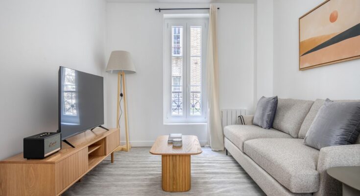 Un salon moderne et lumineux aménagé avec un canapé gris, une télévision sur meuble en bois et une fenêtre haute.
