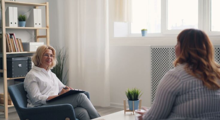 Une thérapeute en blouse blanche écoute attentivement sa patiente lors d'une consultation en cabinet.